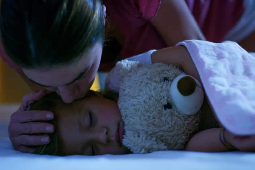 Mother Tucking Child Into Bed And Kissing Sleeping Child With Teddy Bear