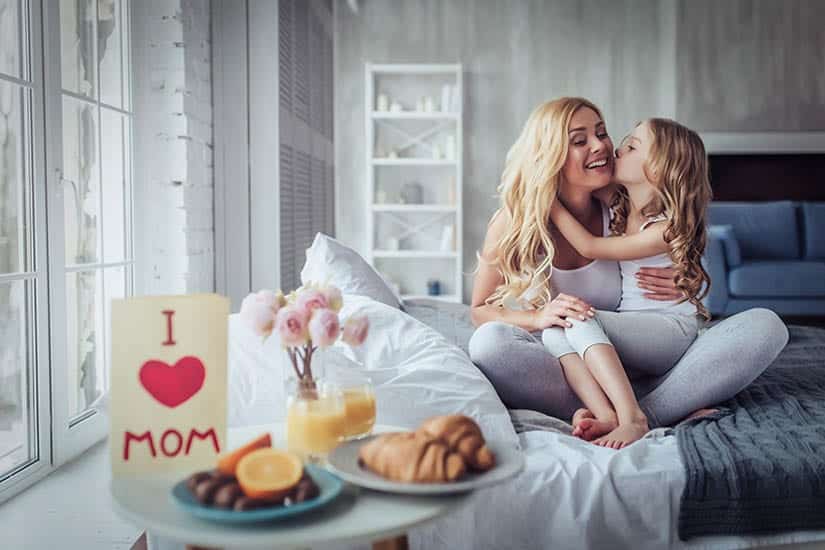 Mother and daughter sitting in bed, daughter kissing mother on cheek. Breakfast on Nightstand with "I love Mom" sign