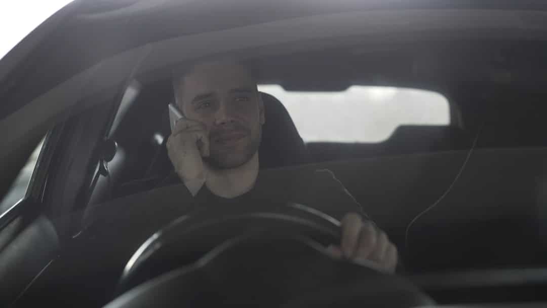 Man sits in the driver's seat, talking on a cellphone and smiling slightly inside a car, steering with one hand.