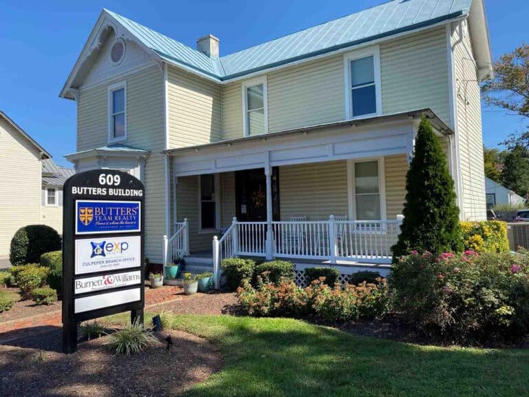 Front view of a two-story beige building with a light blue metal roof and white porch, a sign in the yard reading '609 BUTTERS BUILDING' for a real estate office.