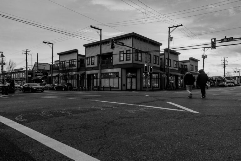 Two people walk across a crosswalk at a busy street corner with a row of storefronts in the background, on a gray urban day.