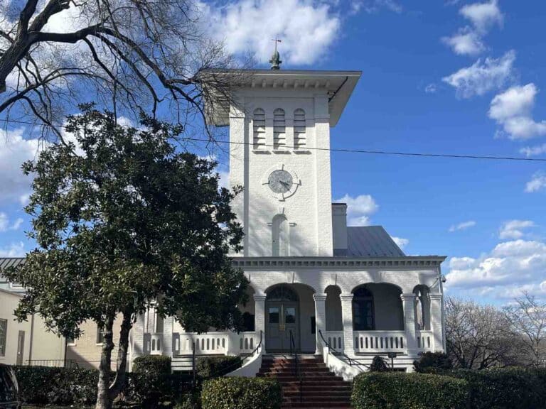 White brick clock-tower building with a wraparound porch and stairs, set against a bright blue sky.
